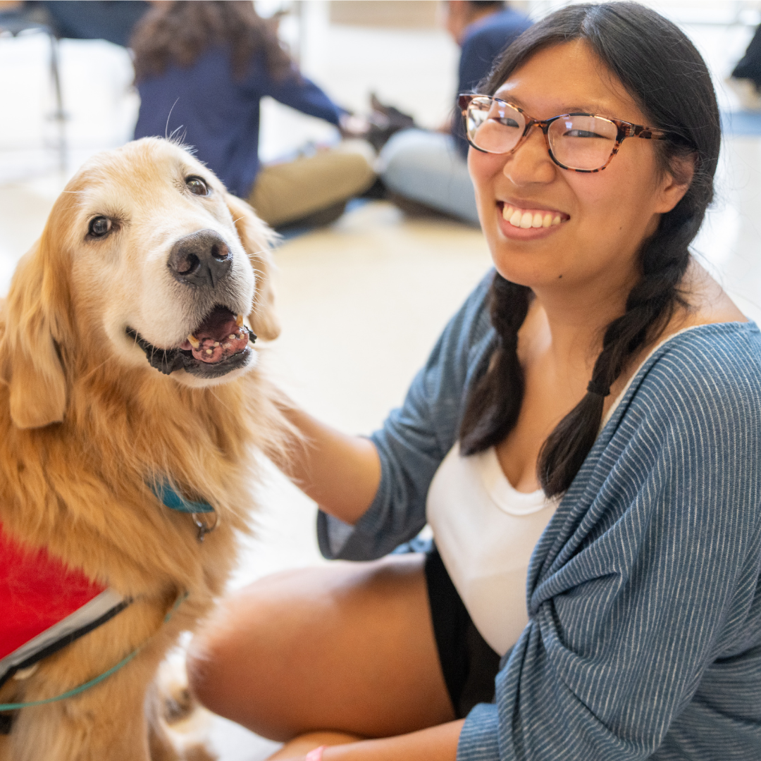 A student with glasses and a big smile sits with and pets a dog.